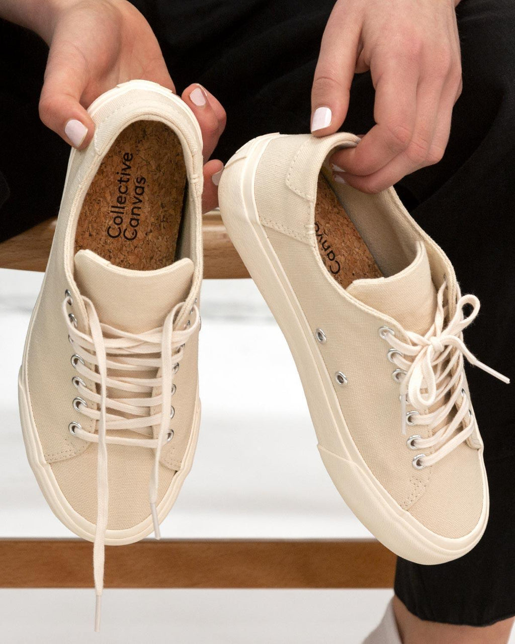 Hands holding a pair of Baan Natural trainers, showing cork insoles and loose laces