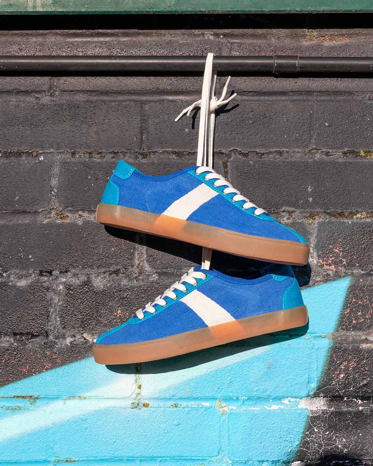 Pair of Looper Hemp Blue trainers hanging by their laces against a painted brick wall