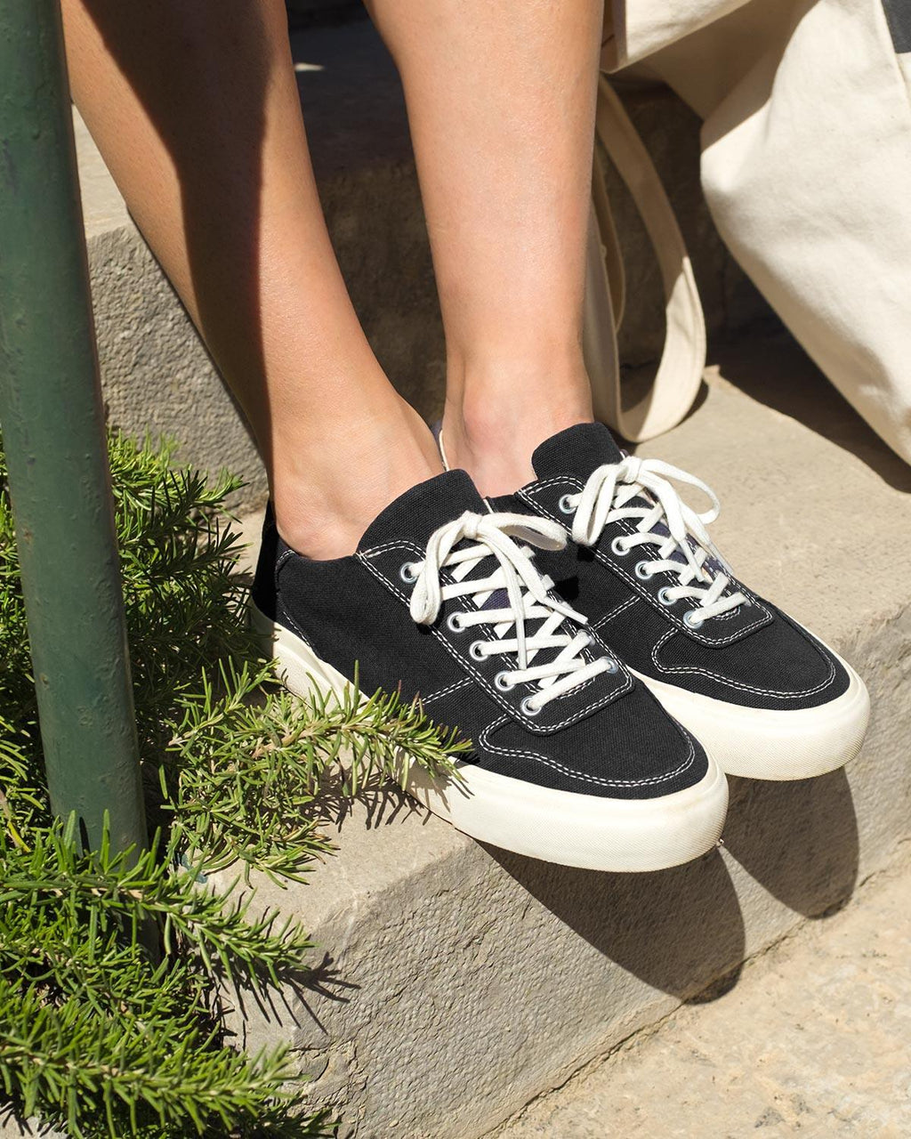 Wearing Bal Black/White trainers seated on stone steps beside a plant and canvas tote bag