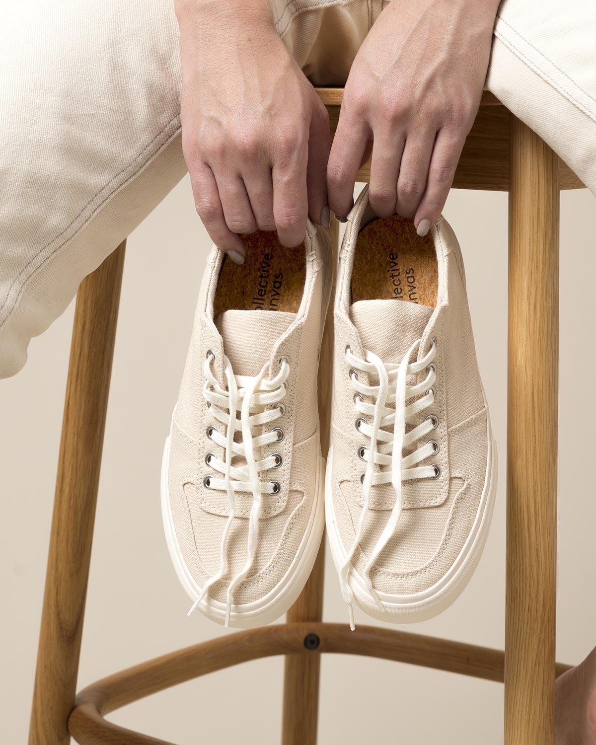 Holding a pair of Bal Natural trainers by the laces while seated on a wooden stool in cream trousers