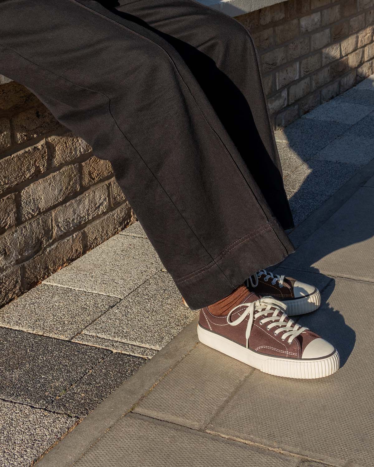 Straat Hemp Coffee trainers worn with rust socks and dark wide-leg trousers, seated on a brick wall